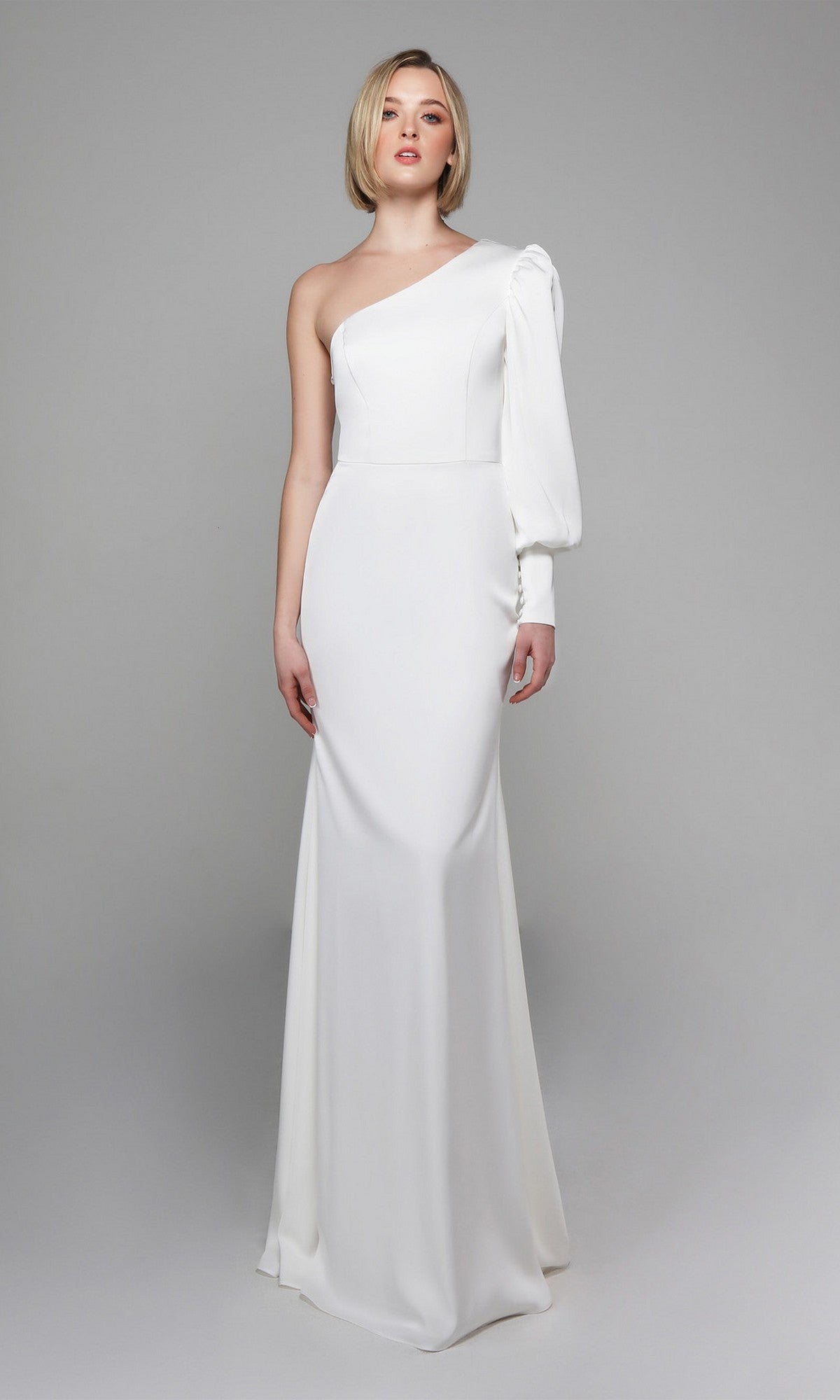 A woman wears the Alyce 7065 Long White Dress, a fitted one-shoulder evening gown with an asymmetrical sleeve, set against a plain gray background.